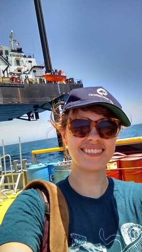 A photo of science writer Monica Kortsha in sunglasses and hat with a raised boat in the background. 